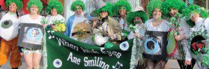 People in a line smiling with green wigs and st Patricks day items holding a green banner that say when Irish eyes are smiling