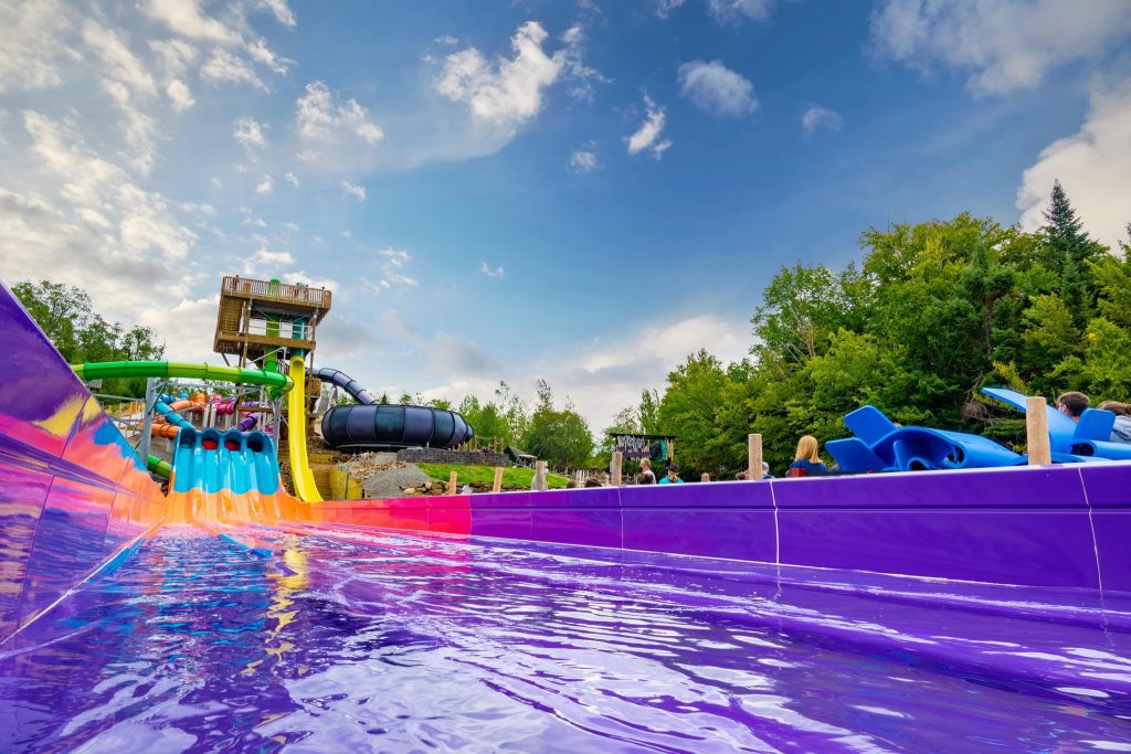 Bottom of multi colored water slide with water collecting at base an people walking up side with mats