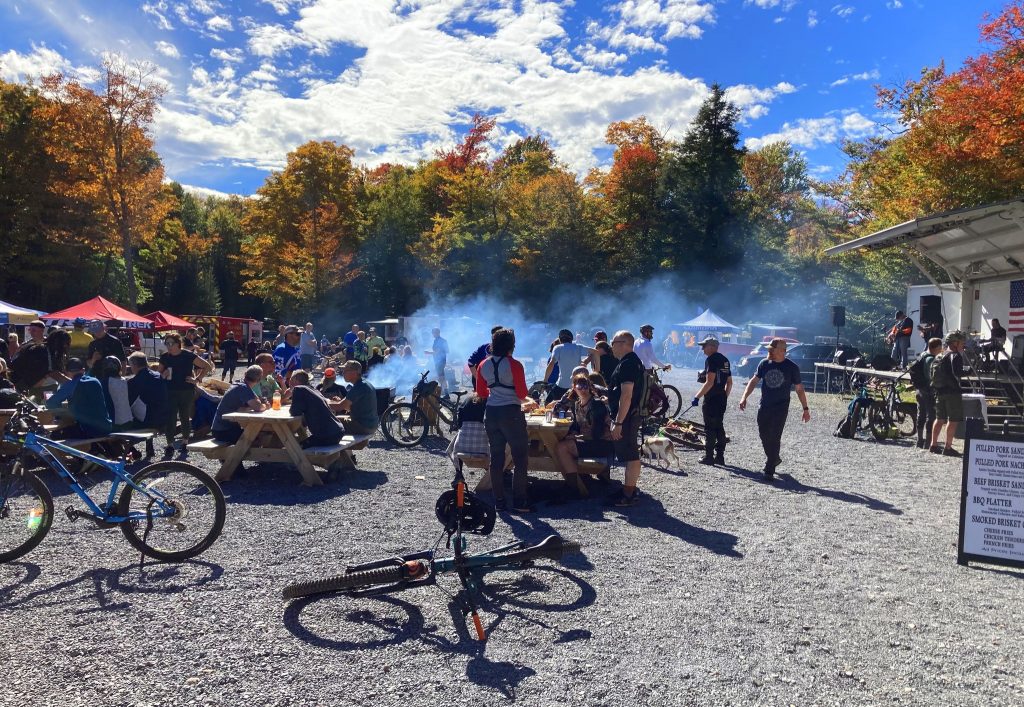 Crowd gathered at an outdoor festival with picnic tables, bicycles, food vendors, and autumn foliage in the background.