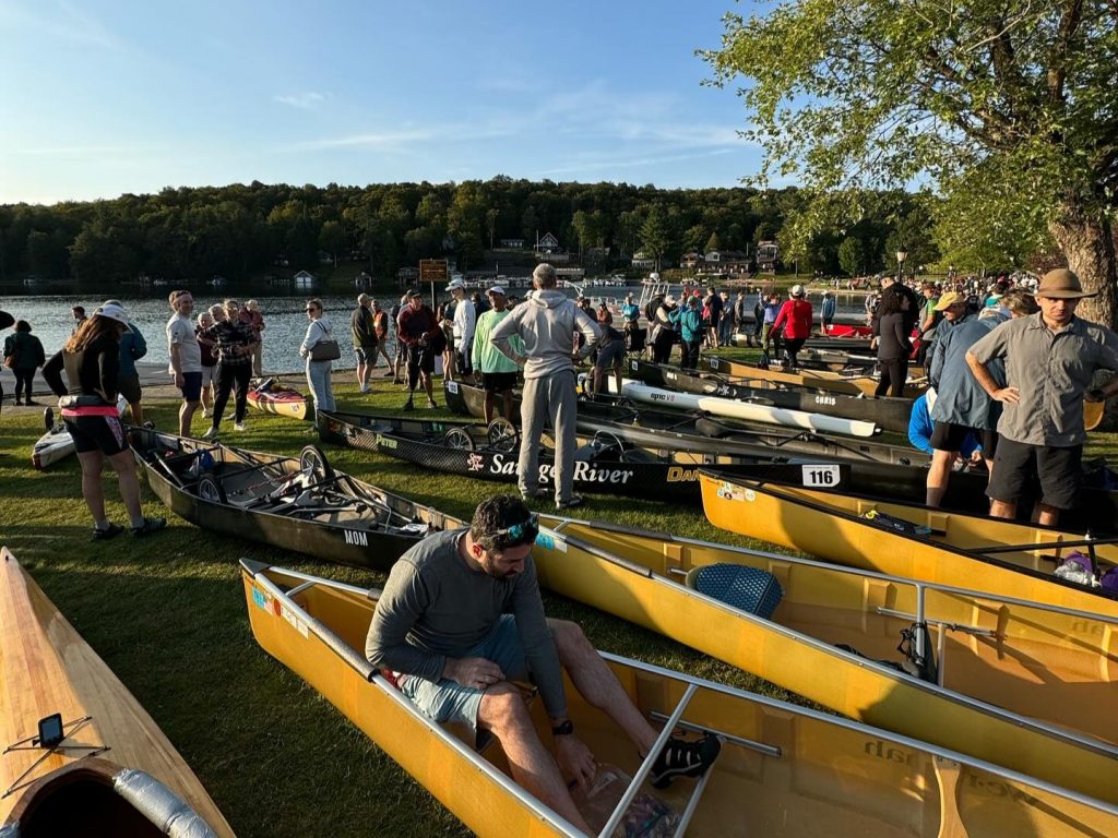 Man sits in boat before race with boats and people standing in background