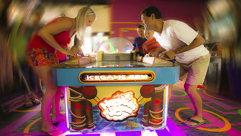 Two adults playing arcade games against themselves and kids watching them