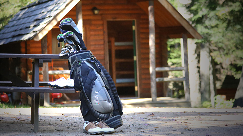 A cabin in the background with golf clubs leaning against a picnic table and gold shoes sitting around the clubs in the front of the image