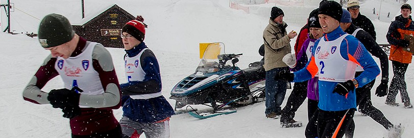 People dressed in racing gear running in the winter past a cabin and a snowmobile
